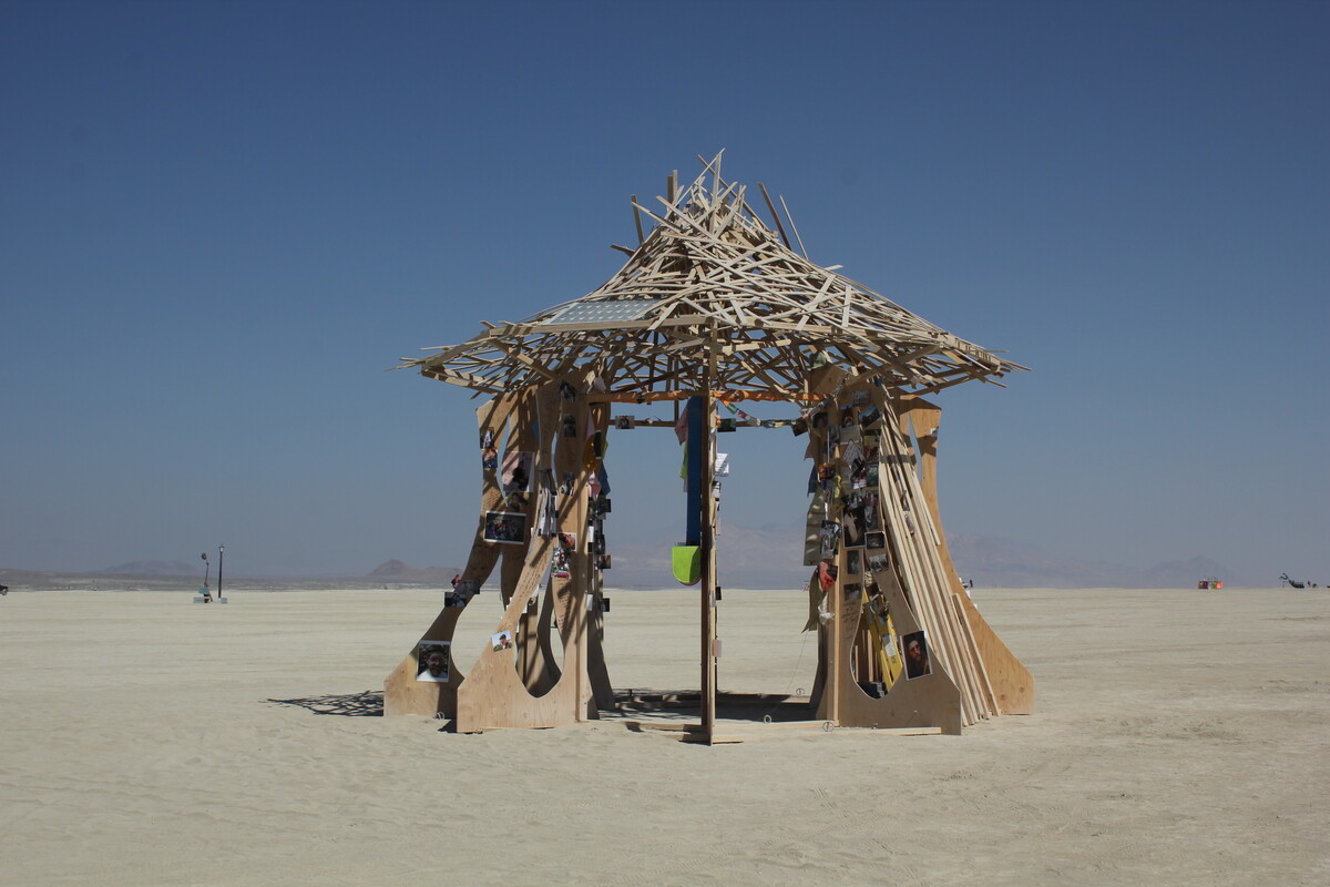 The Temple of Greg standing alone on the Burning Man playa under a clear blue sky — a wooden gazebo with curved plywood walls, photos and mementos pinned to its pillars