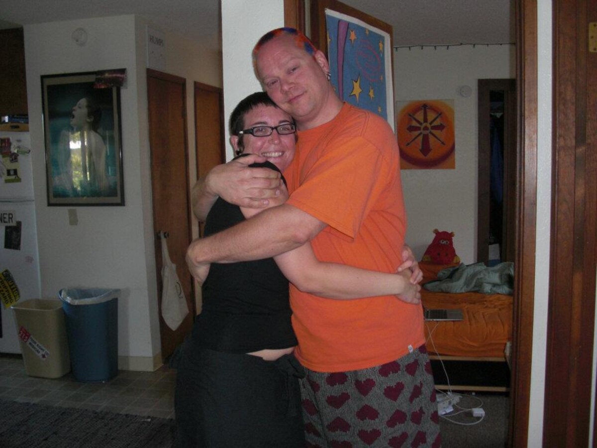 Rob in an orange t-shirt and heart-patterned shorts hugging Erin in a black top and glasses, both grinning, in his SLO apartment hallway. A red stuffed hippo sits on the bed visible through the doorway behind them.