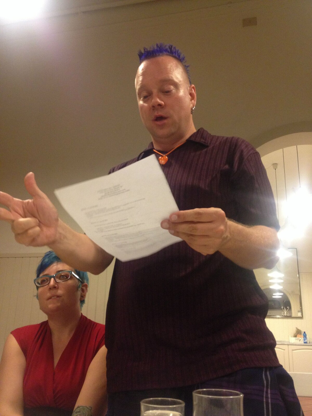 Rob with a blue-purple mohawk in a dark button-down shirt, standing at a restaurant table reading from a paper and gesturing mid-sentence. Erin with bright blue hair, red top, and glasses sits beside him, looking up.