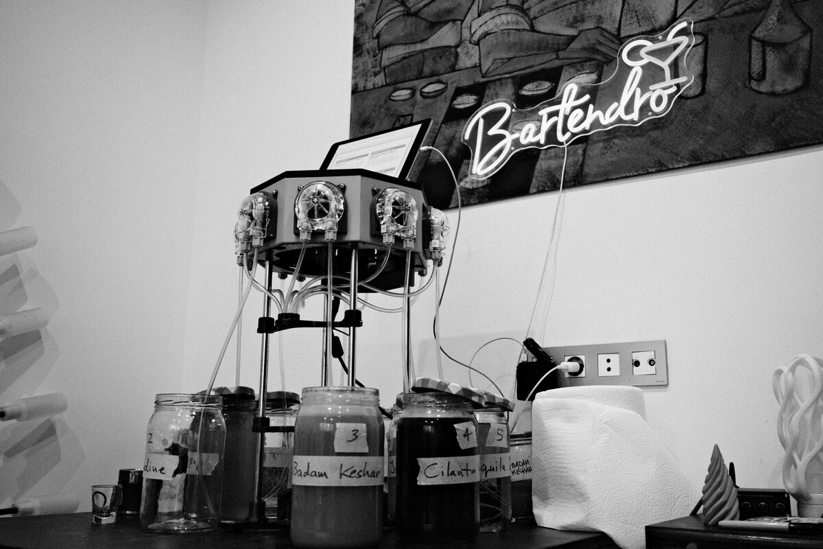 The Bartendro cocktail robot with its glass orbs and tubing, positioned beneath a glowing neon Bartendro sign, in black and white.