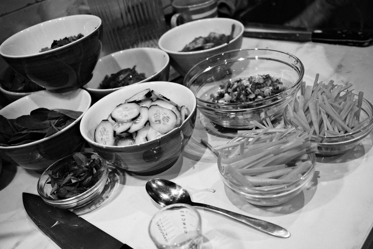 Overhead view of a food spread with bowls of sliced cucumbers, green beans, salads, and tapas arranged on a counter, in black and white.