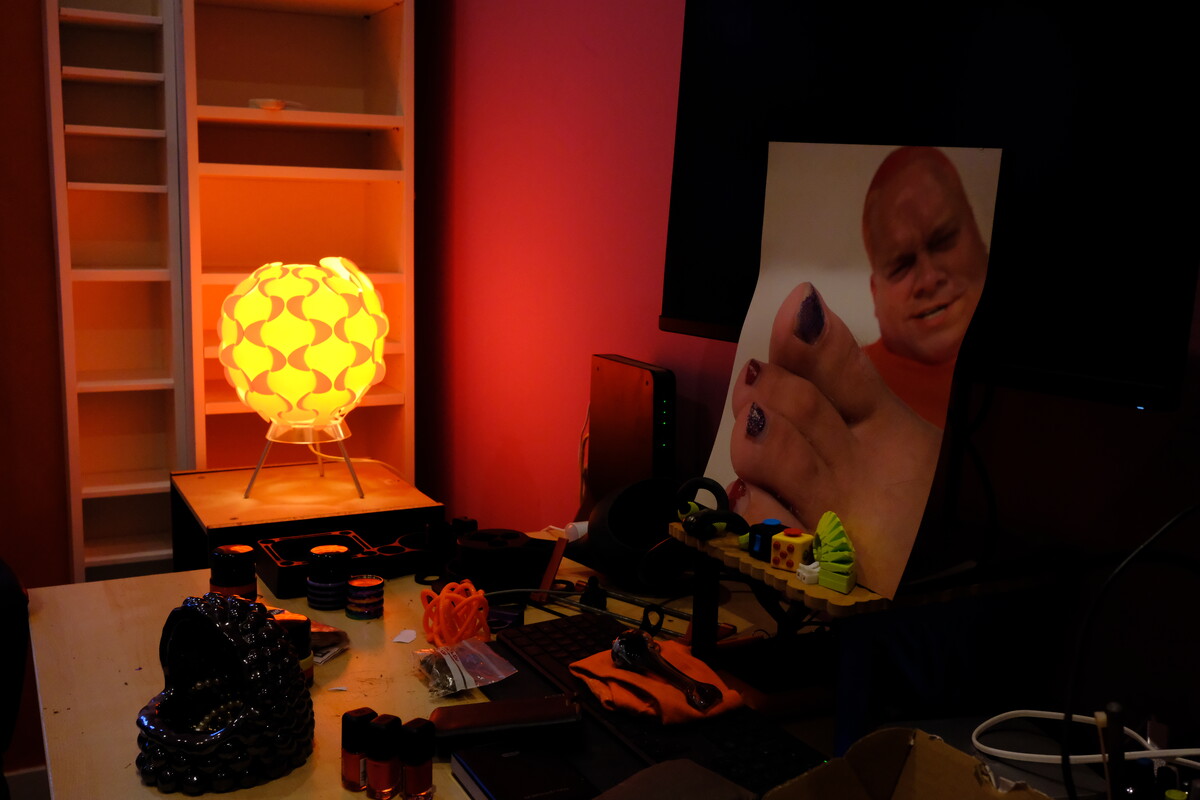 Rob&rsquo;s portrait glowing on a screen beside a warm globe lamp and scattered objects in his red-lit apartment.