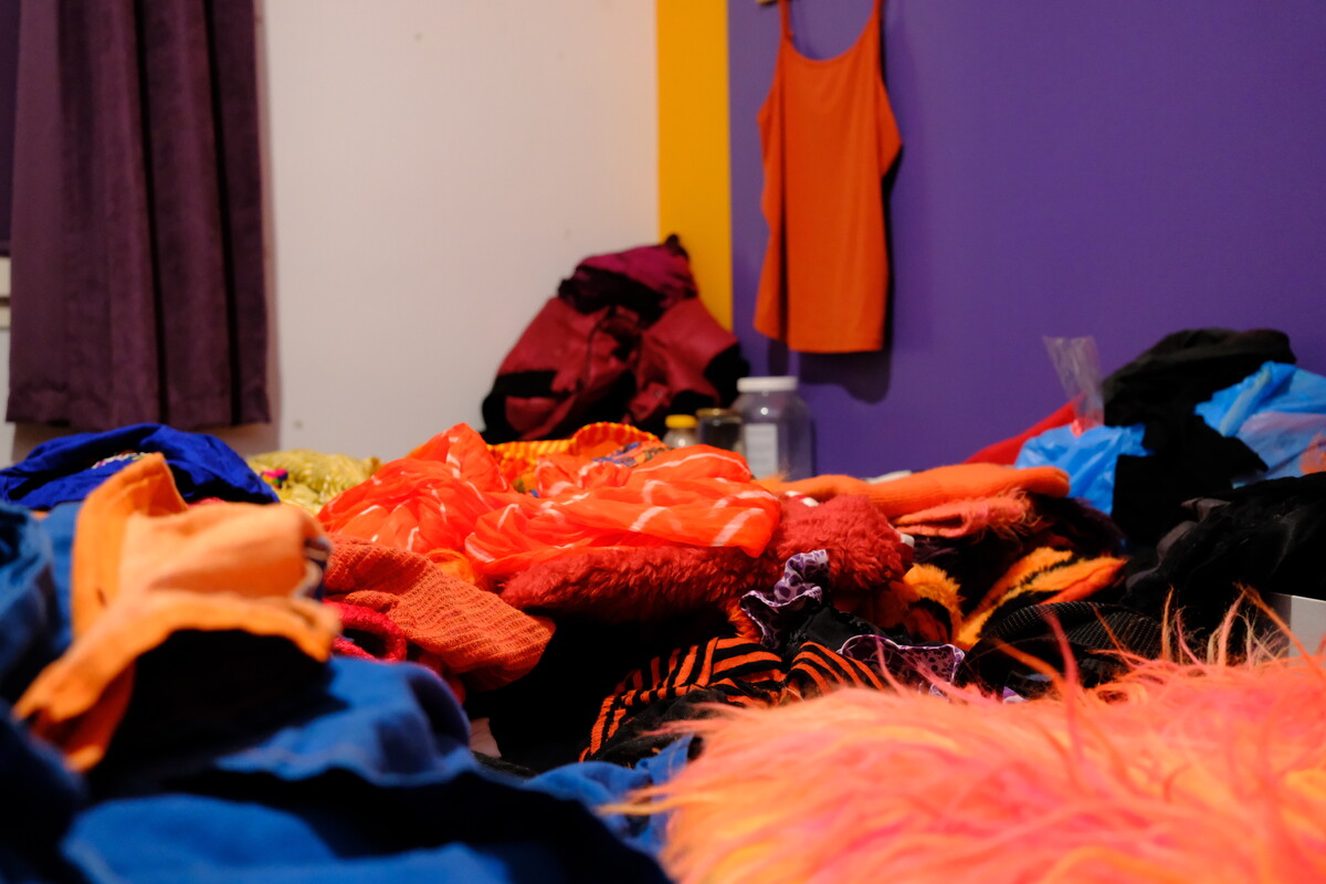 A bed heaped with costumes in orange, blue, magenta, and faux fur against a purple and yellow wall.