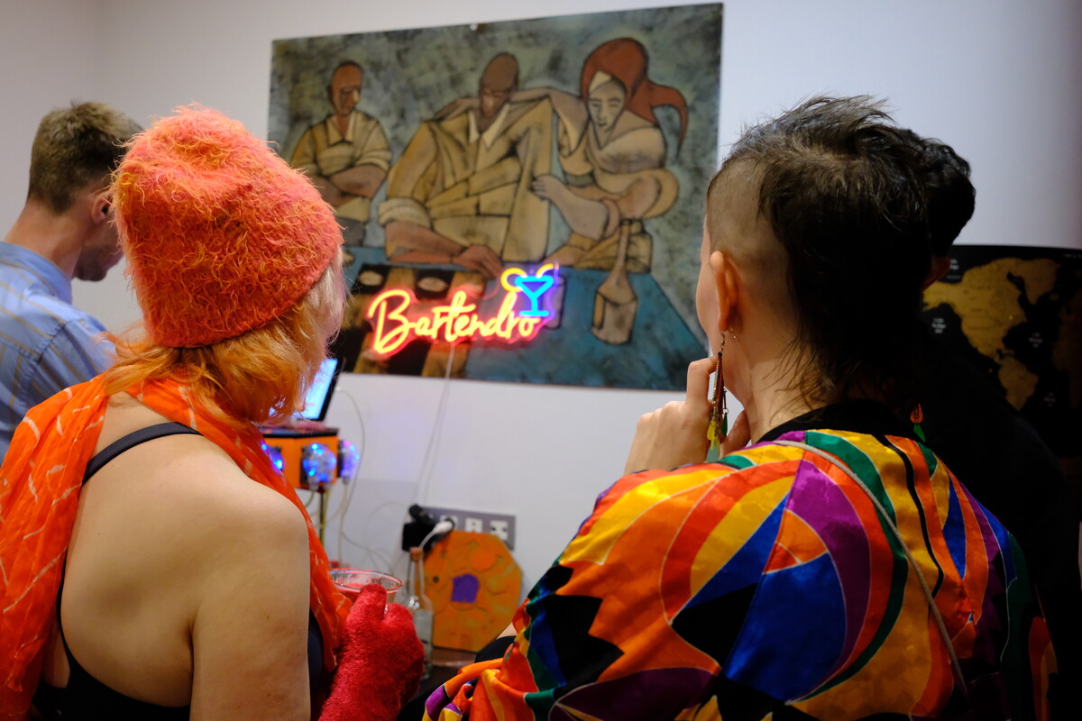 Three friends seen from behind, standing before the Bartendro neon sign and a large painting, one wearing an orange fuzzy hat, another in a colorful silk shirt.