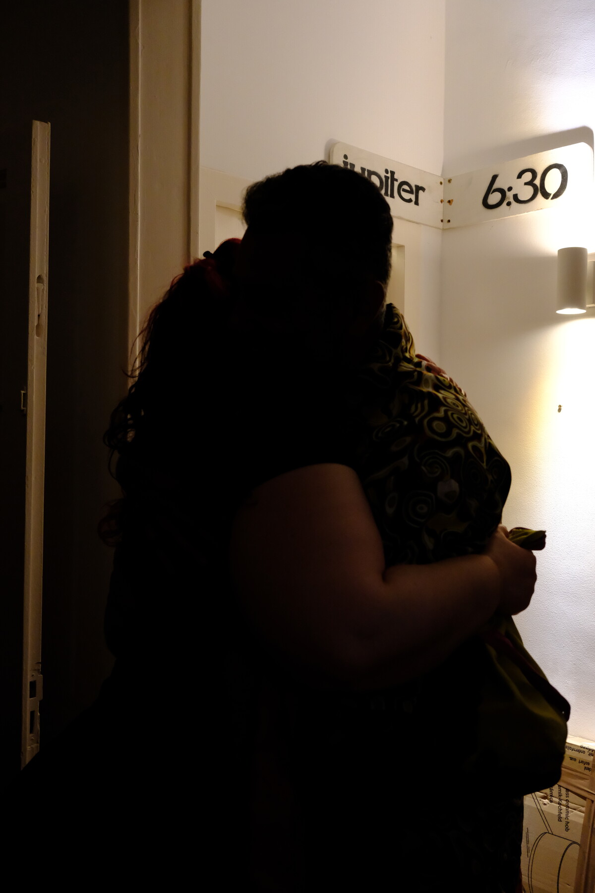 Two silhouetted figures embracing in a hallway beneath a &ldquo;Jupiter 6:30&rdquo; sign, lit by a single warm wall sconce.