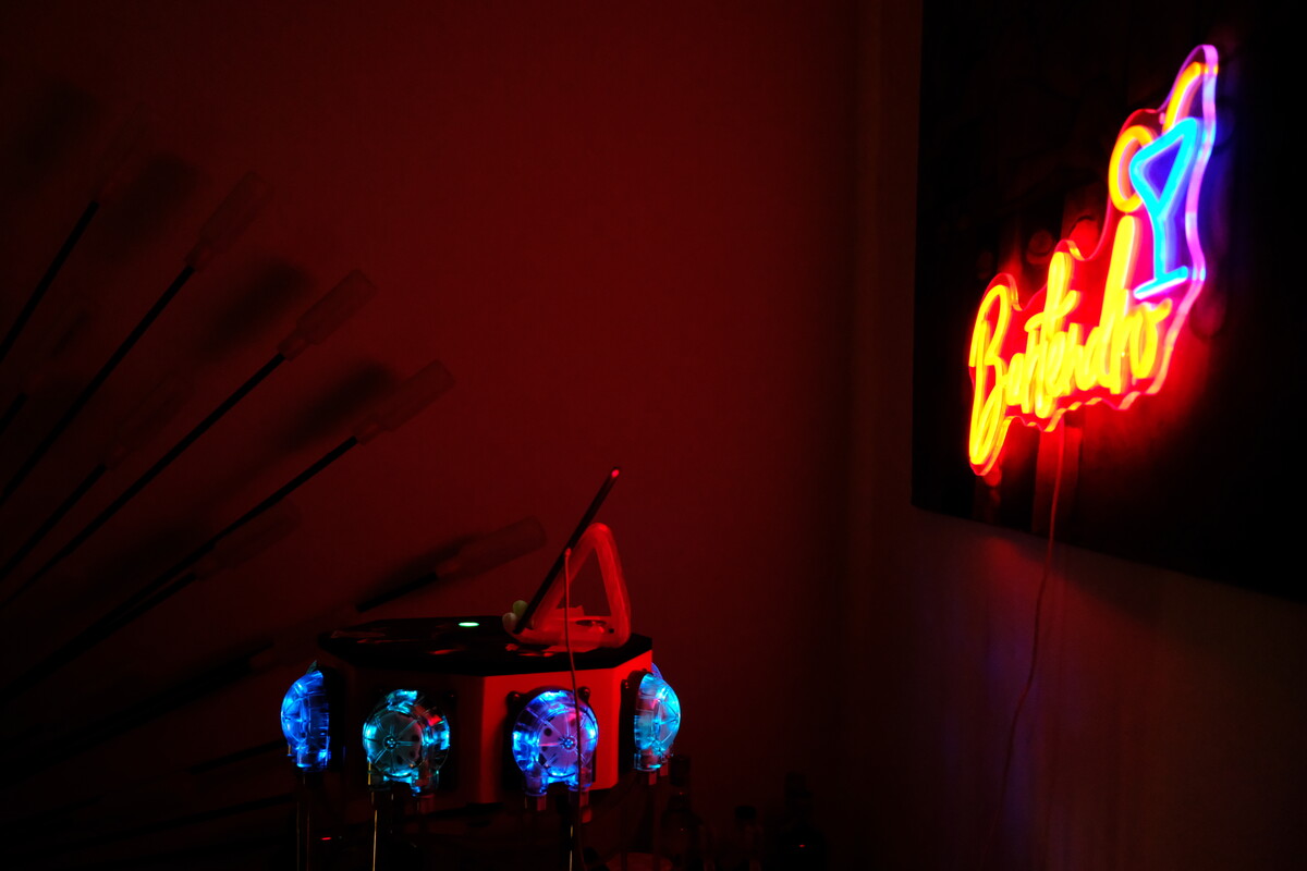The Bartendro robot glowing blue in darkness, with the neon Bartendro sign and cocktail glass lit in red, yellow, and blue on the wall behind.