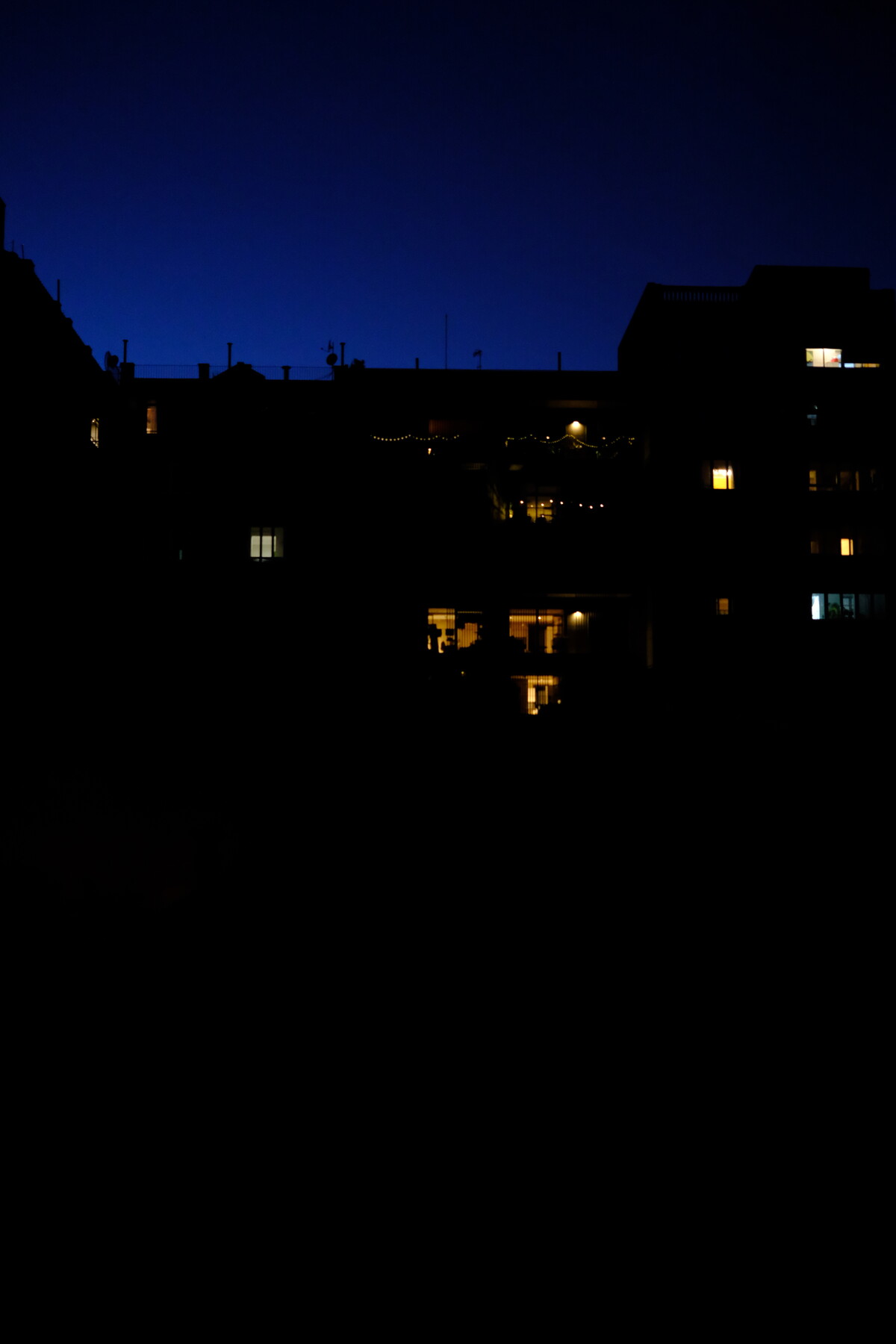 Barcelona rooftops at night, dark silhouettes against a deep blue twilight sky, with fairy lights strung on a distant terrace and warm windows glowing.