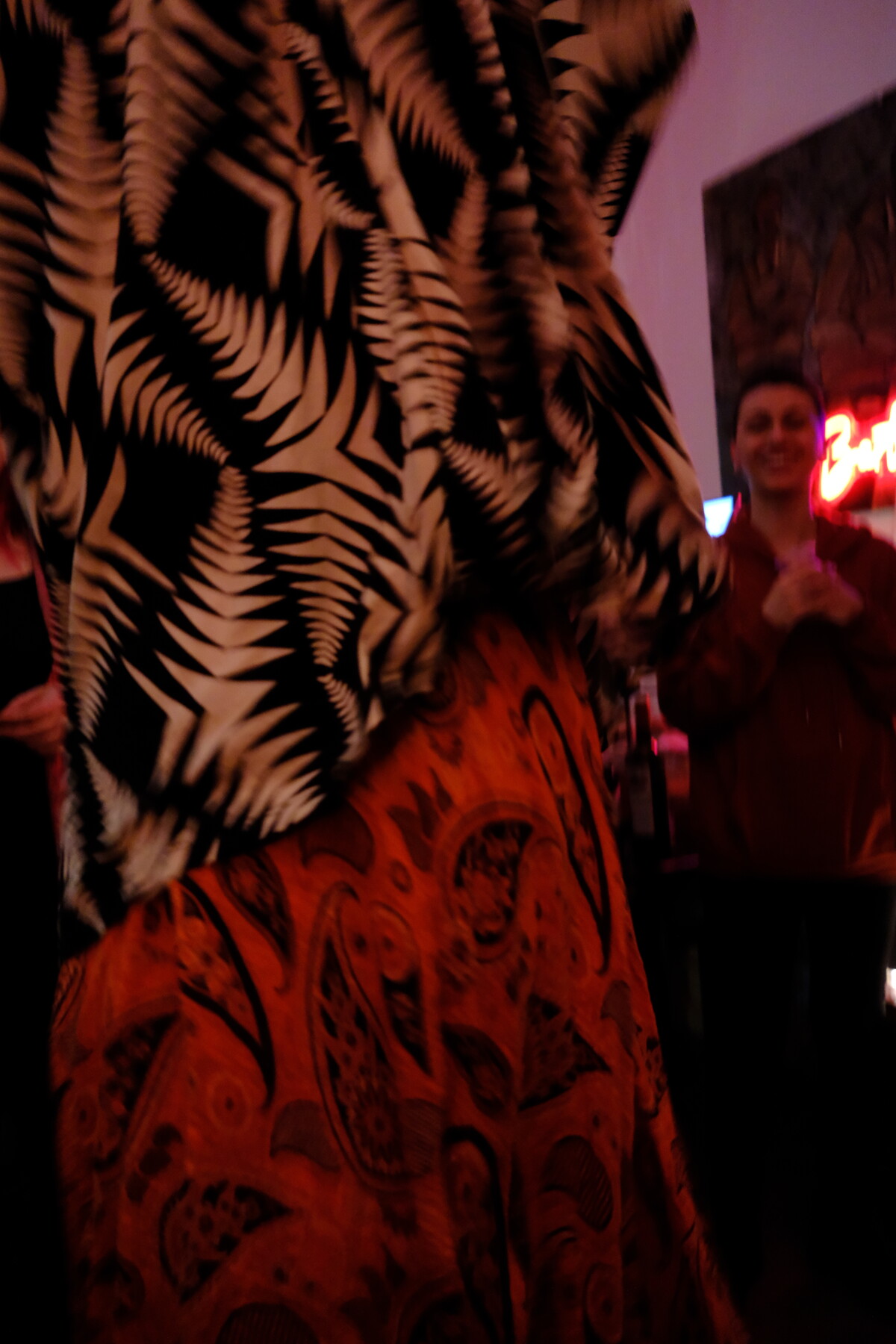 Close-up of layered fabrics in motion &mdash; a black-and-gold fern-patterned wrap over a red paisley skirt, blurred figures and warm light behind.