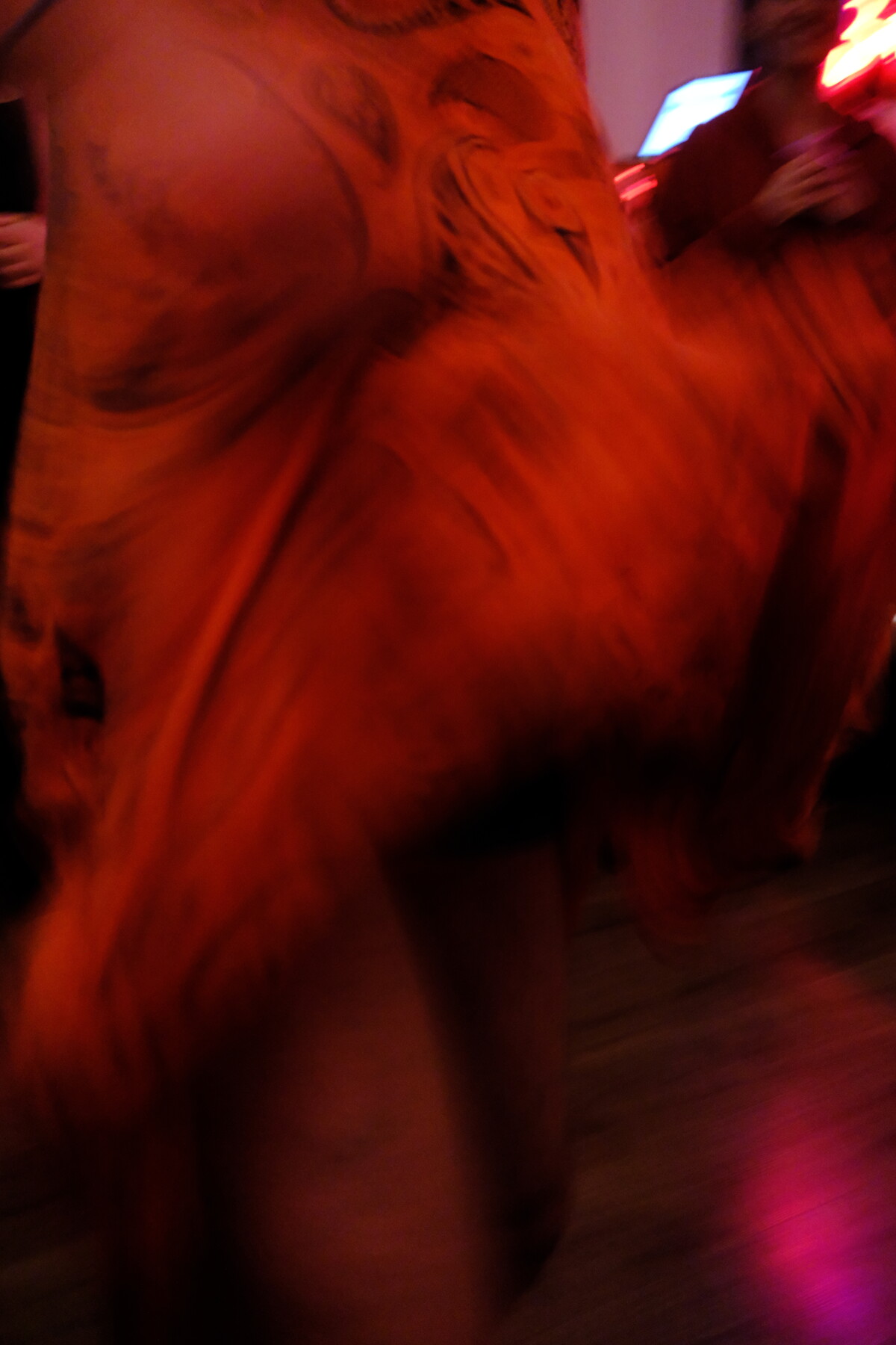 Abstract motion blur of swirling red and orange fabric or movement, with a neon sign faintly visible in the background.