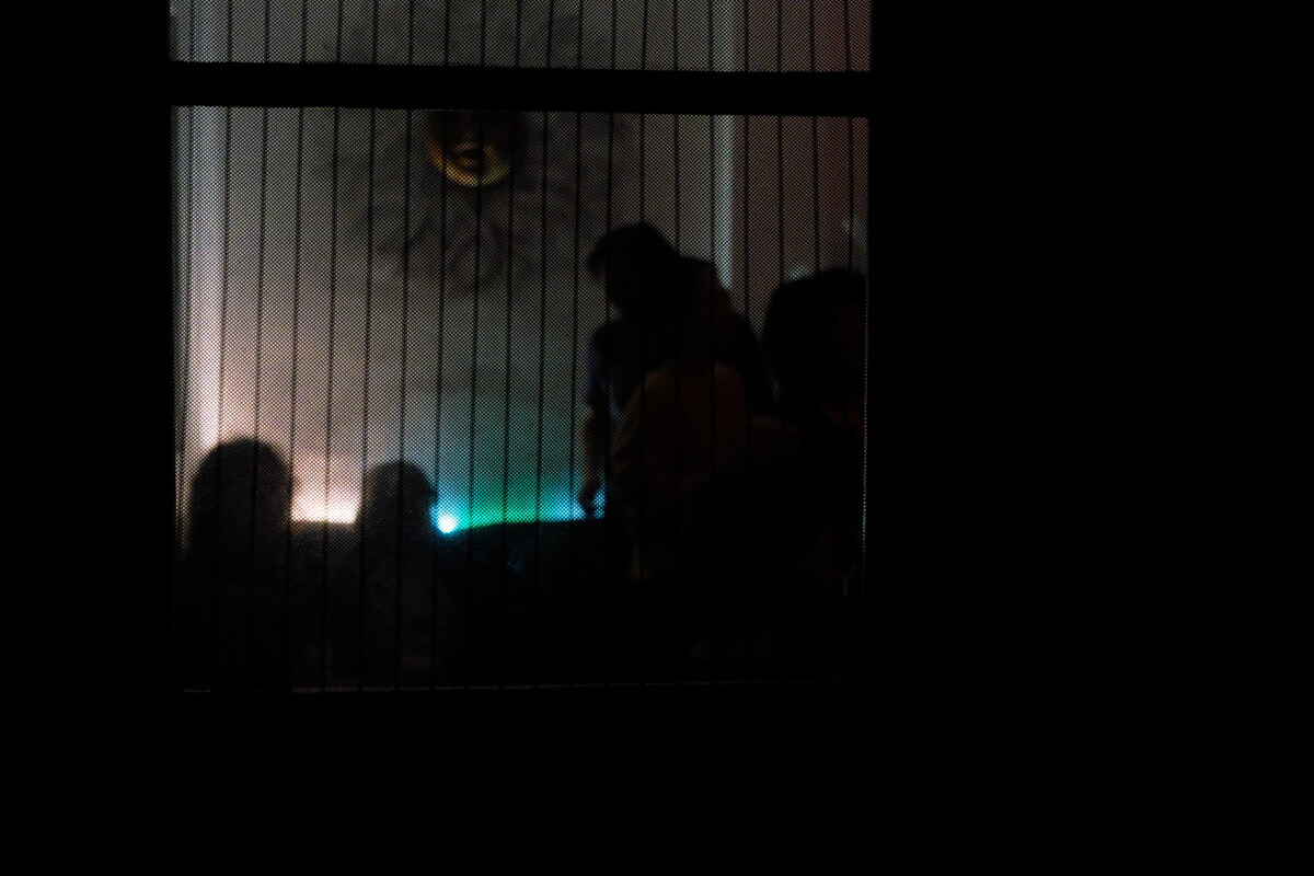 View from outside through slatted shutters into a lit room, silhouettes of people gathered inside, a faint green glow among them.