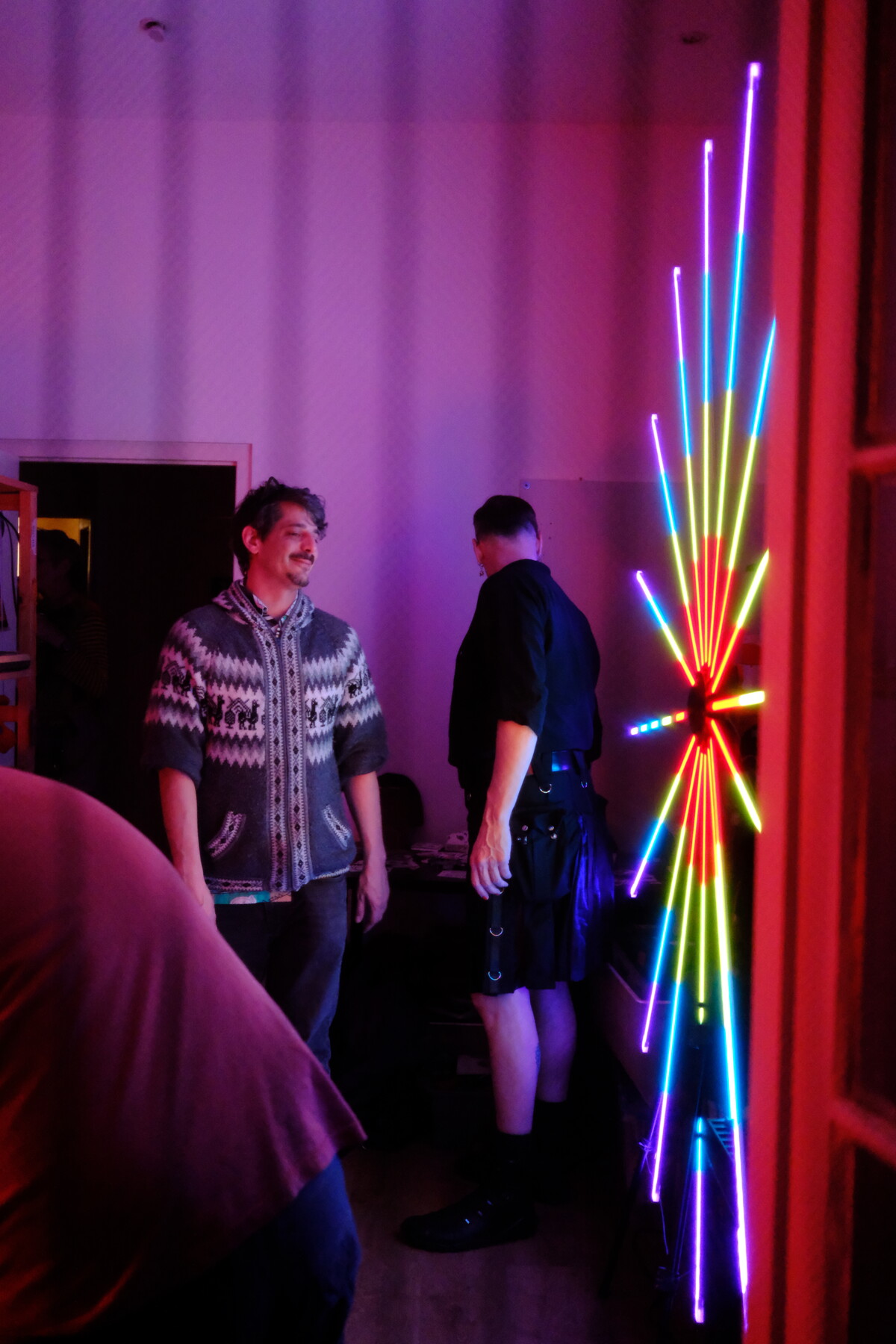 A full-spectrum rainbow LED starburst installation glowing cyan, yellow, green, red, and magenta, with two partygoers standing nearby.