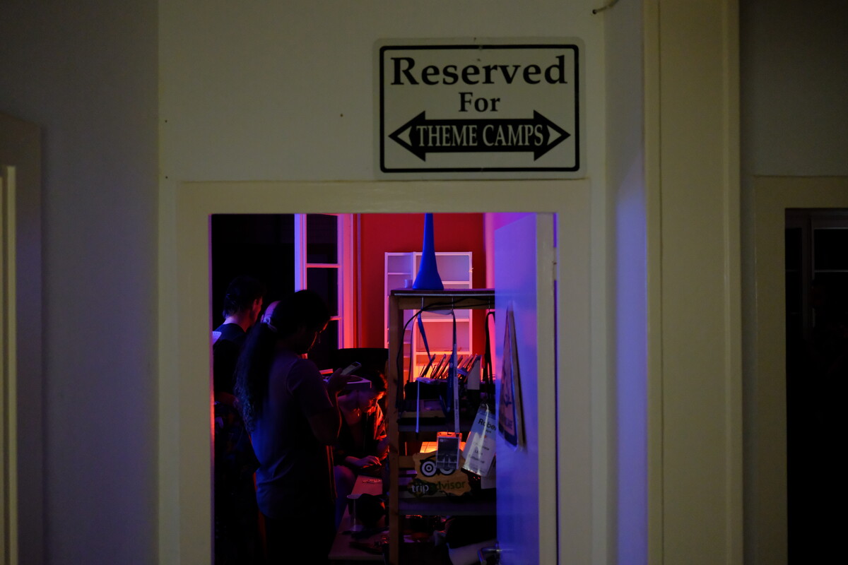 A sign reading &ldquo;Reserved For Theme Camps&rdquo; mounted above a doorway, with red and purple light spilling through from the room beyond.