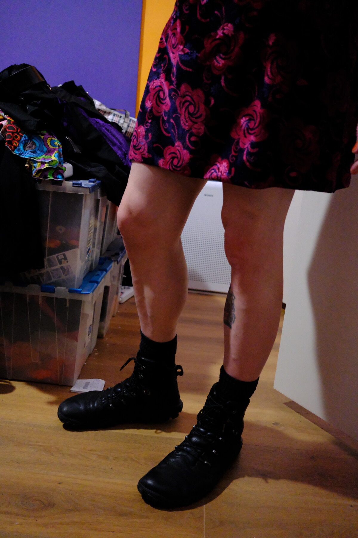 Lower body shot of someone wearing black combat boots, a magenta baroque-print skirt, and a colorful headscarf, standing against a purple and yellow wall.