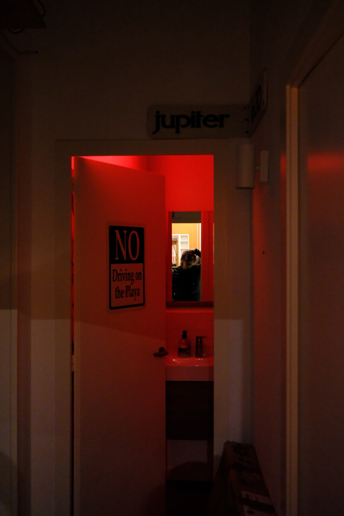 A hallway bathed in red light, the word &ldquo;jupiter&rdquo; mounted above a doorway. Inside, a Burning Man sign reads &ldquo;NO Driving on the Playa,&rdquo; a mirror reflects a figure, and toiletries line the sink.