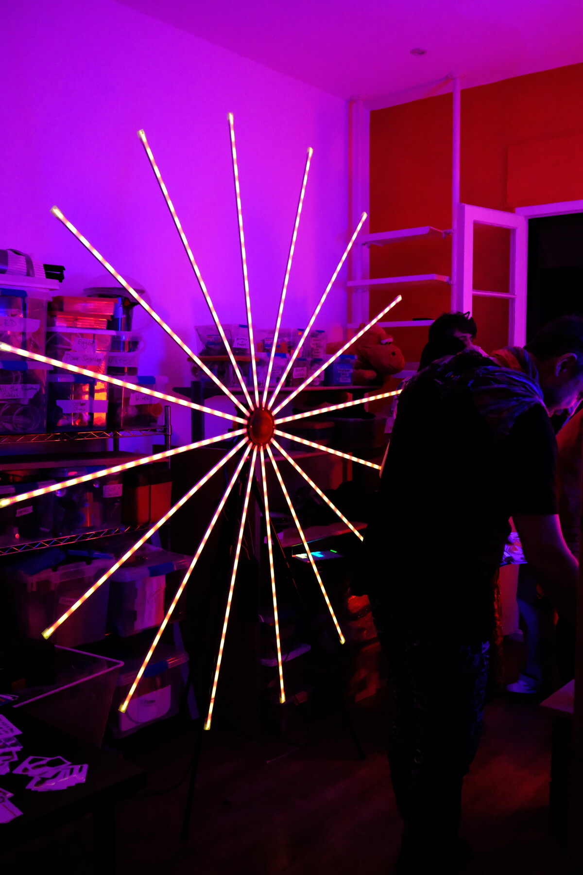 A starburst LED sculpture radiating from a central point, its rods glowing in warm colors against purple and red light. A silhouetted figure stands behind it.