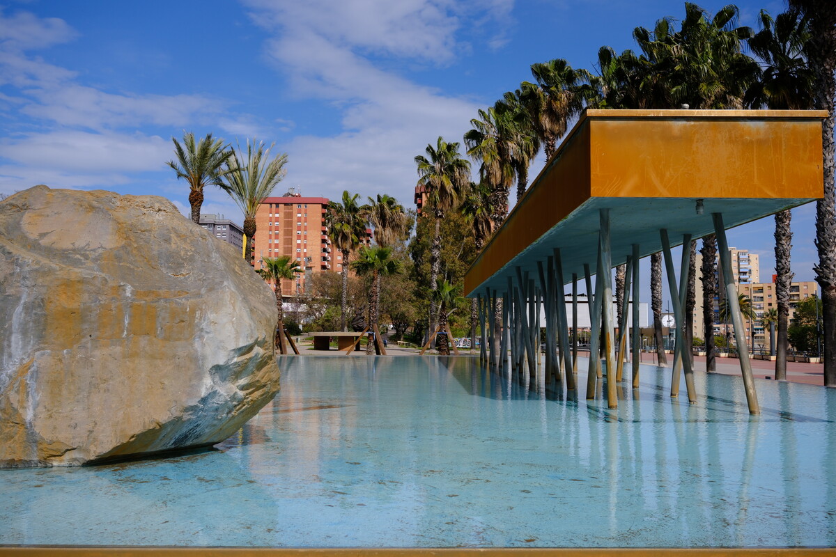 A Barcelona park with a shallow reflecting pool, palm trees, a large boulder, and a modern canopy structure. Apartment towers in the background under a partly cloudy sky.