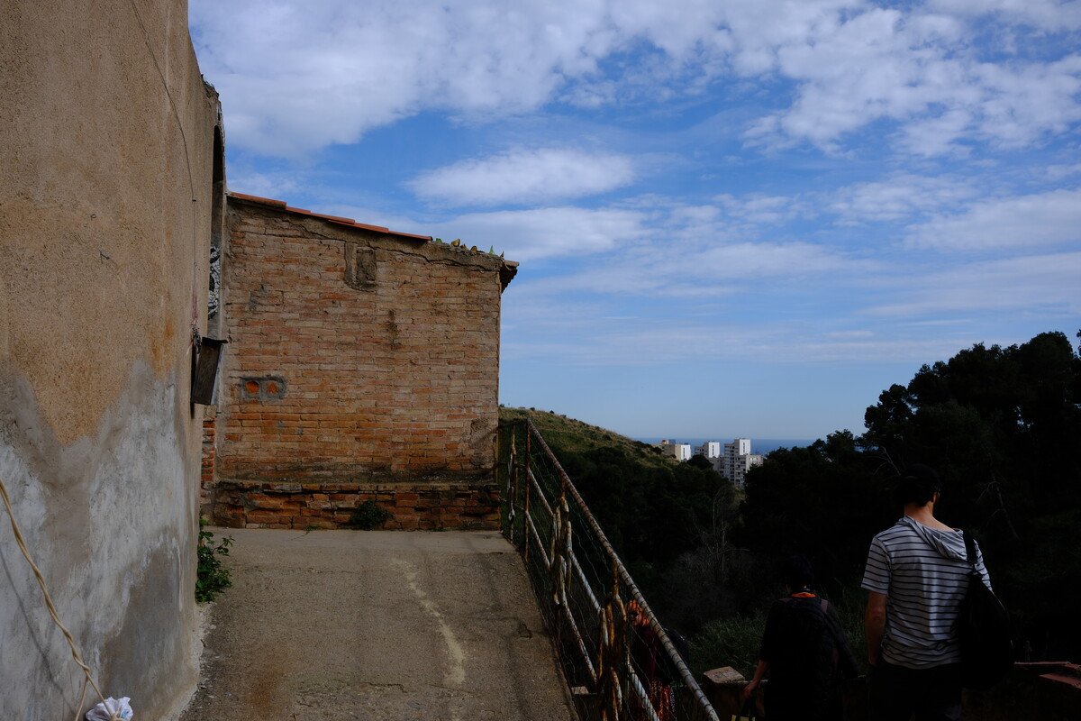 A narrow hillside path between old brick buildings and a metal railing, with a view across trees to distant towers and the sea. A person walks ahead in a striped shirt.