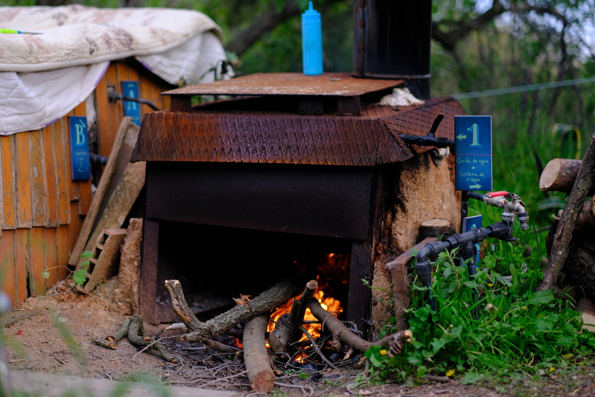 A rustic outdoor wood-burning stove made from brick and reclaimed metal, with an active fire burning inside, in a garden setting.