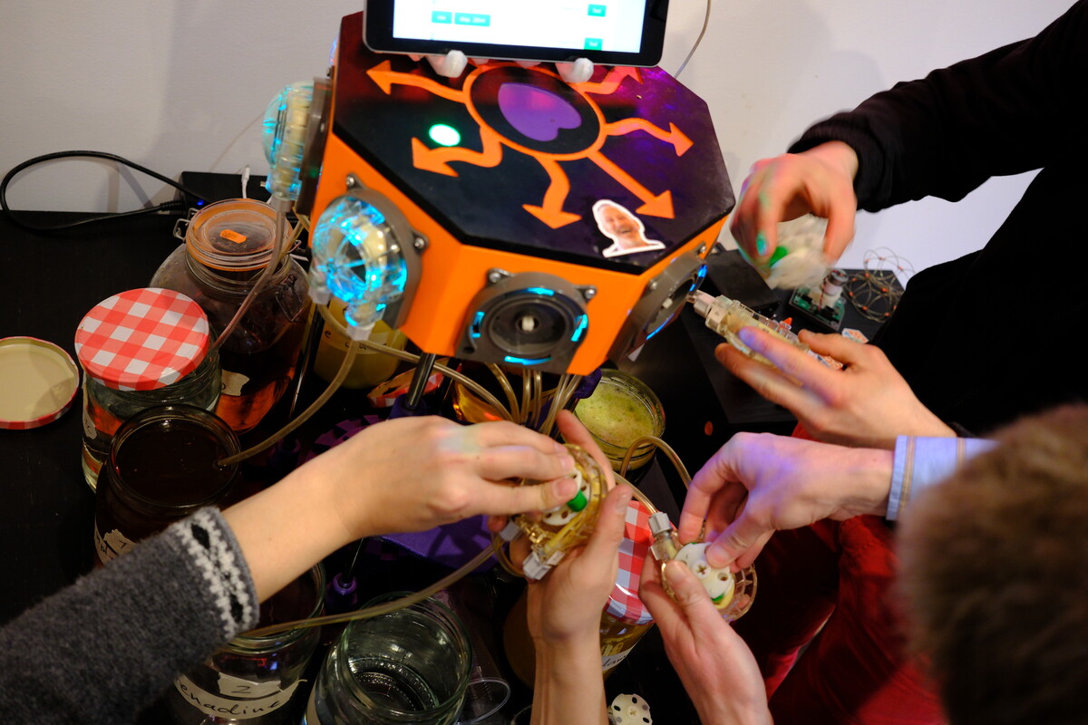 Close-up of the Bartendro cocktail robot &mdash; an orange and purple hexagonal body with LED lights and an iPad mounted on top &mdash; surrounded by hands reaching in with mismatched jars and glasses.