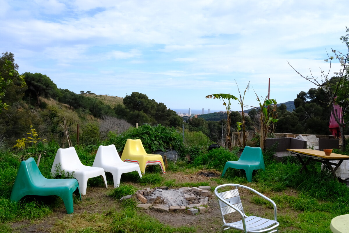Colorful plastic chairs &mdash; green, white, yellow, turquoise &mdash; arranged in a circle around a fire pit in an overgrown hillside garden, with the Barcelona skyline visible in the distance.