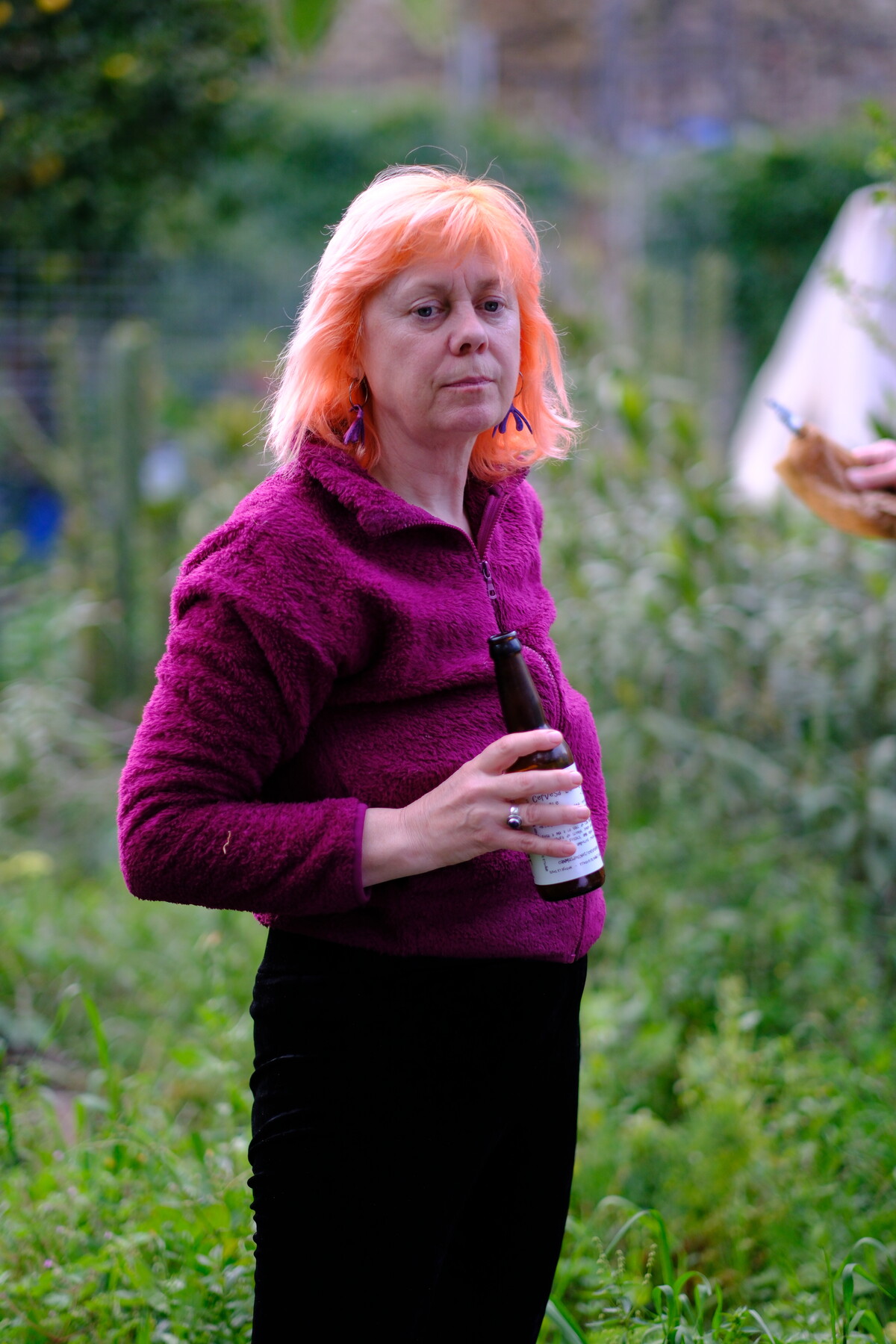 Kirsten standing in a green garden holding a bottle, wearing a purple sweater, her hair bright orange against the foliage.