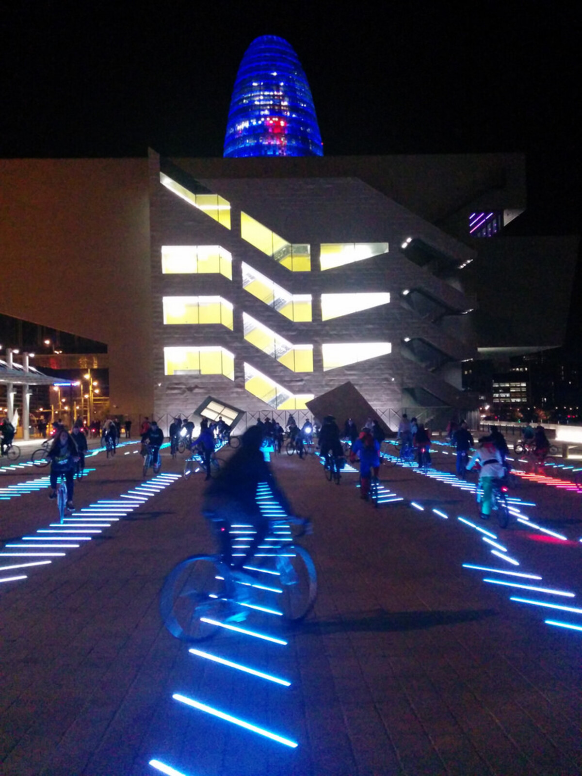 Dozens of cyclists riding at night past the illuminated Museu del Disseny building, with the glowing blue Torre Agbar tower rising behind it, neon blue light strips embedded in the ground casting streaks across the plaza.