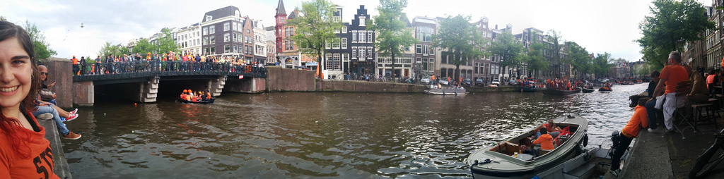 Wide panoramic shot of an Amsterdam canal during King&rsquo;s Day, bridges packed with crowds in orange, boats filling the water, buildings lining both sides. A woman in an orange &lsquo;Start Your Revolution&rsquo; t-shirt is visible at the far left edge.