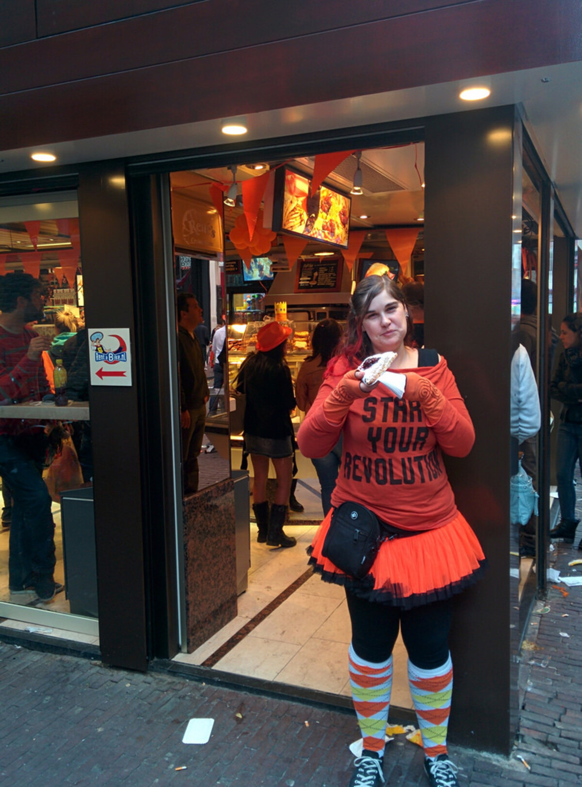 Woman standing in the doorway of an Amsterdam shop during Koningsdag, wearing an orange &lsquo;Start Your Revolution&rsquo; shirt, neon orange tutu over black leggings, colorful knee-high socks, and eating a pastry, with orange decorations and crowds visible inside.