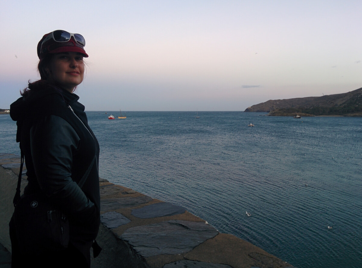 Woman in a dark jacket with sunglasses perched on her cap, standing on a stone seawall at twilight, looking out over a calm Mediterranean bay with boats and a headland in the distance.