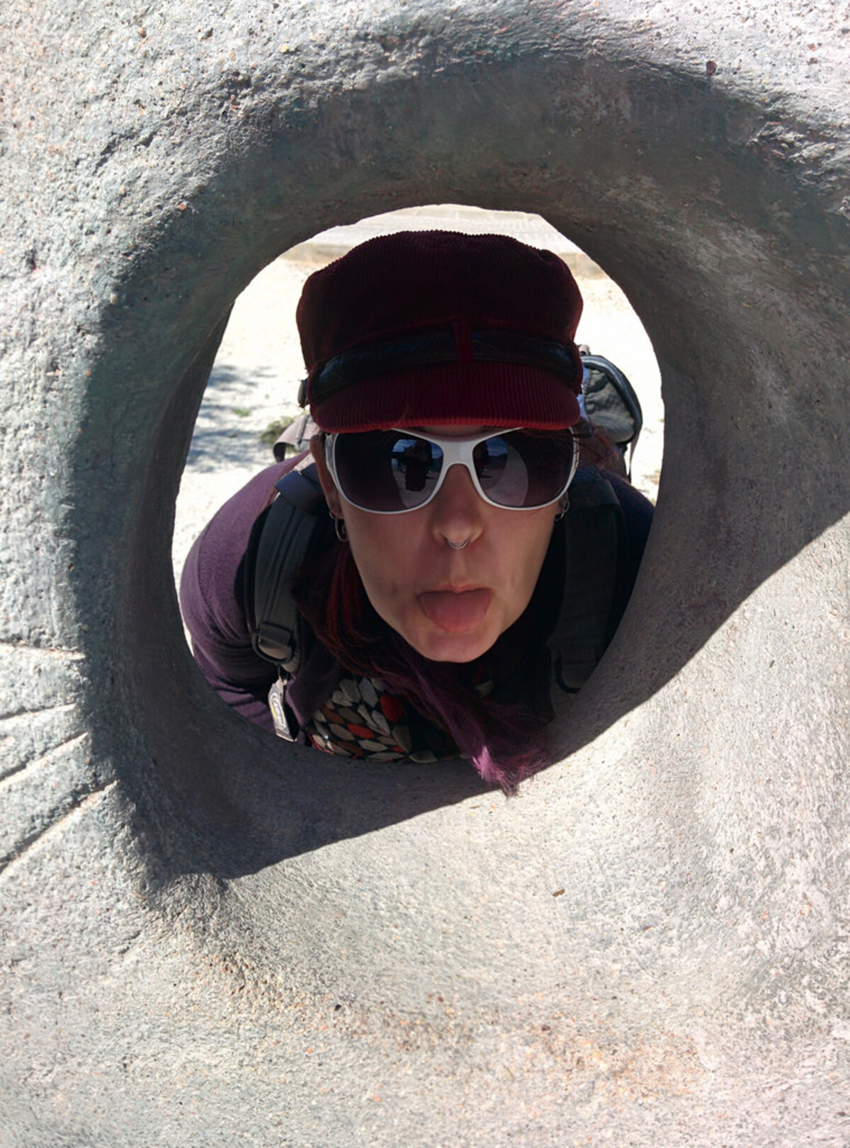 Woman with purple-tinted hair, white sunglasses, and a maroon cap, sticking her tongue out while peering through a circular hole in a large stone sculpture, backpack straps visible.