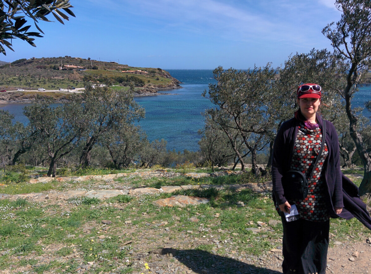 Woman in a colorful patterned top and dark cardigan, standing on a hillside among olive trees overlooking a turquoise Mediterranean inlet with green headlands on either side.