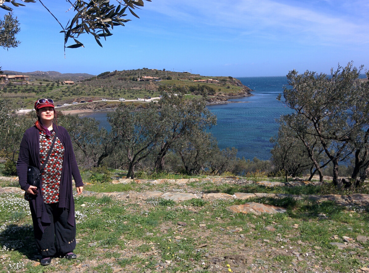 Woman looking up through olive branches at the sky, wearing the same patterned top and dark cardigan, the Mediterranean bay and headland visible behind her on a bright spring day.
