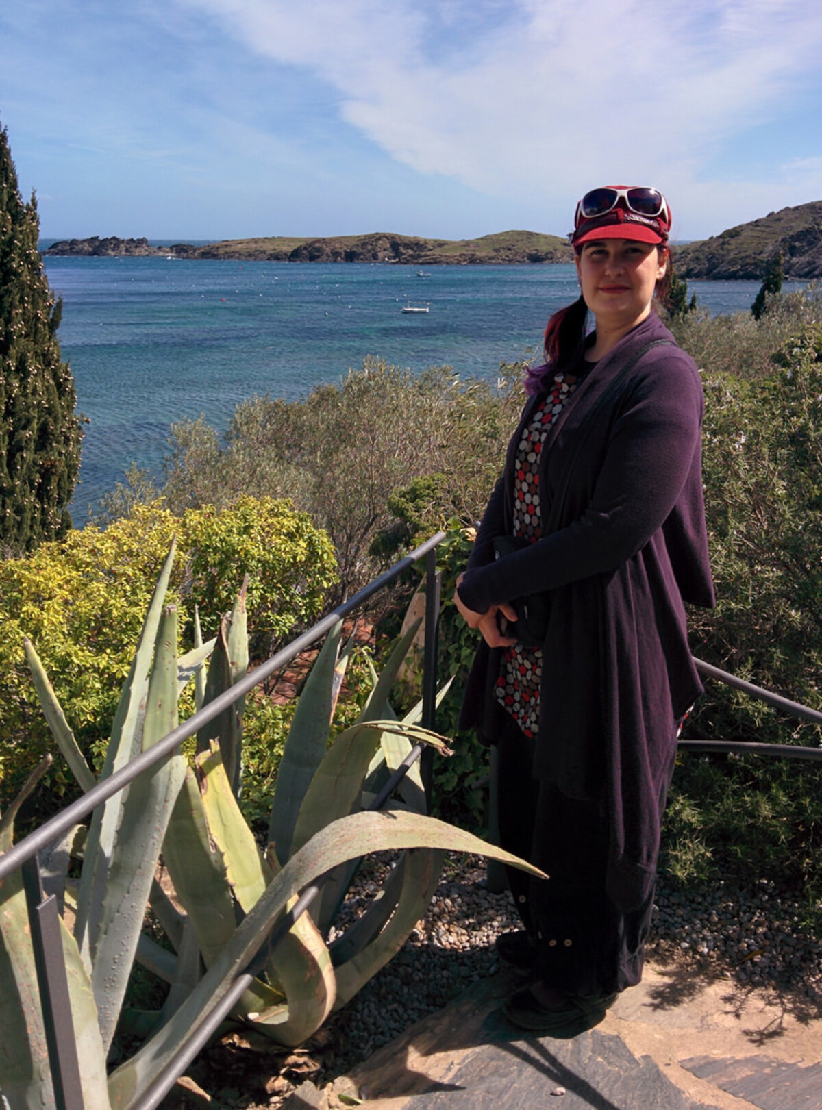 Woman in a purple cardigan and patterned top, standing on stone steps beside a metal railing, with agave plants, cypress trees, and flowering shrubs framing a view of turquoise Mediterranean water and rocky headlands.