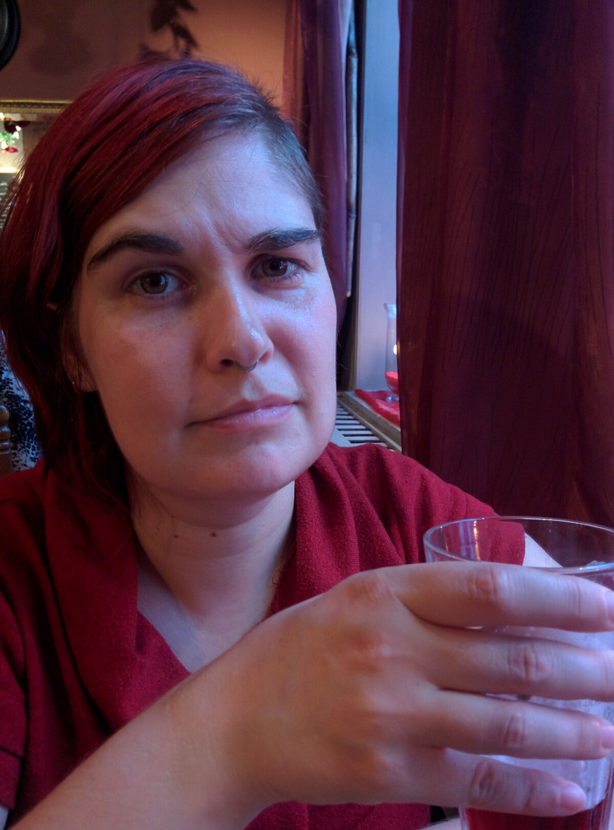 Close-up of a woman with red-tinted hair, holding a glass and looking directly at the camera with a calm, steady expression, dark red curtains and warm bar lighting behind her.