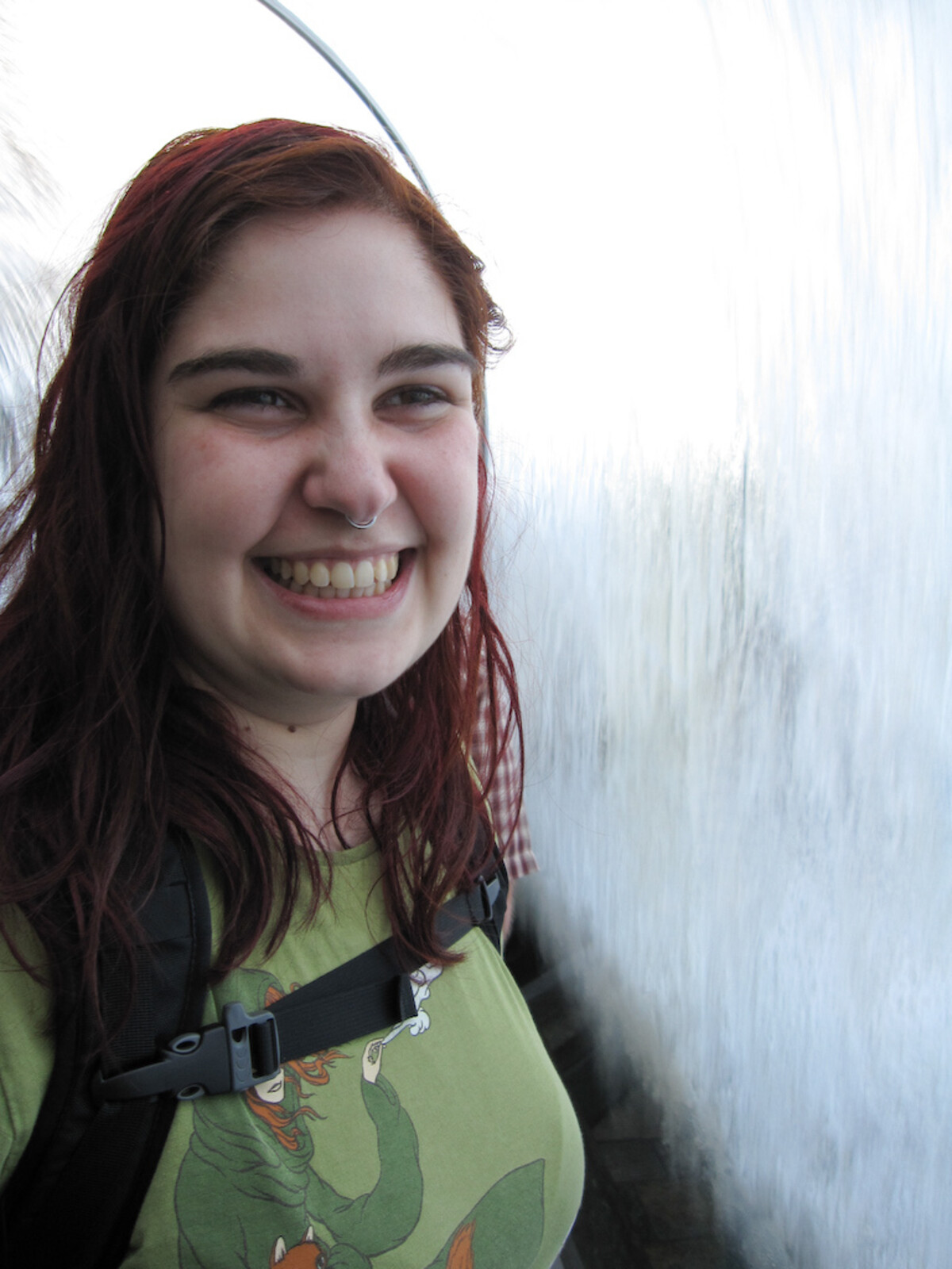 Close-up portrait of a young woman with dark reddish hair, nose ring, and green dragon t-shirt, beaming at the camera with water streaming down glass behind her at an aquarium.