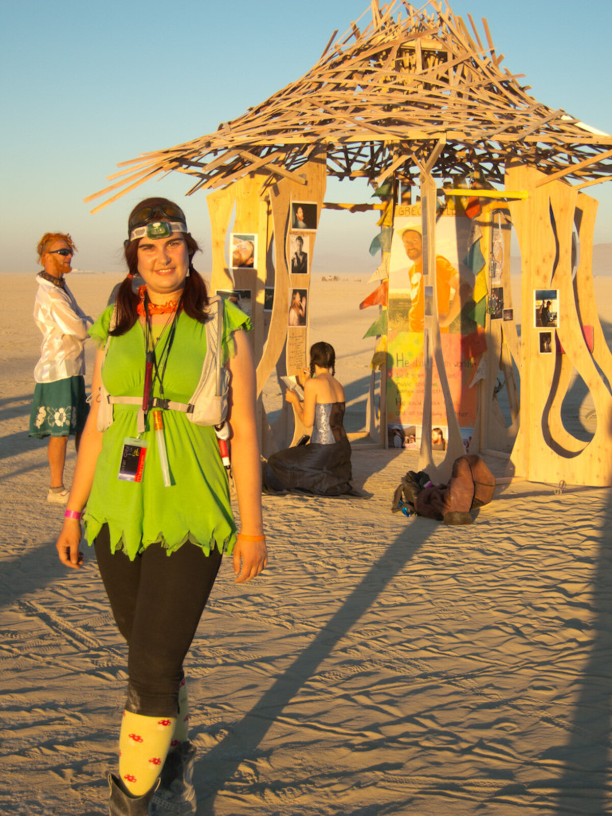 Woman in a green fairy-cut top with yellow socks and boots, standing on playa sand at sunset in front of a wooden memorial temple covered in photos and tributes at Burning Man. Gizmo stands in the background admiring the temple, while a woman crouches to write a message on the temple structure.