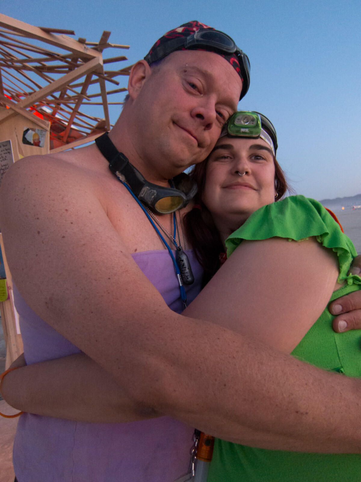 A couple embracing at Burning Man at dusk, the man in a lavender tube top with goggles and a flame-print cap, the woman in green with a headlamp, the wooden temple structure behind them.