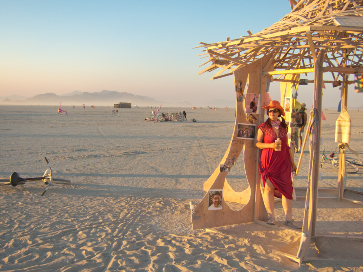 Woman in a red sarong and wide-brimmed red hat, leaning against the wooden memorial temple on the playa, holding a drink, with photos of the deceased pinned to the temple walls and the vast desert behind her.