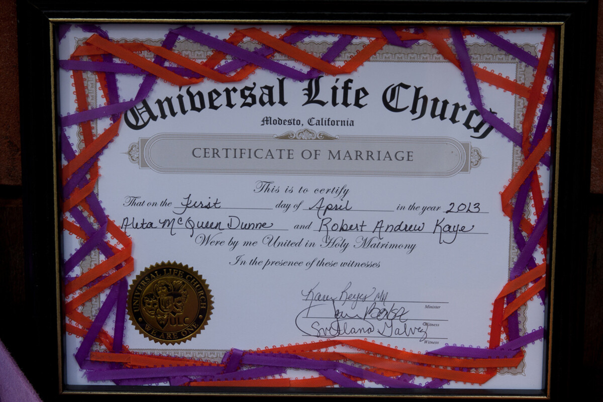 Marriage certificate from the Universal Life Church, now mounted in a black frame with decorative red and purple ribbons woven around the border, displayed on a table among colorful plates.