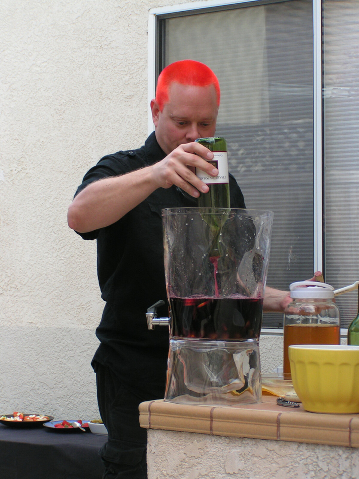 Man with bright neon red-orange buzzcut, wearing a black button-up shirt, pouring wine from a bottle into a large glass beverage dispenser filled with dark sangria on an outdoor patio table, with tapas and a yellow bowl beside him.