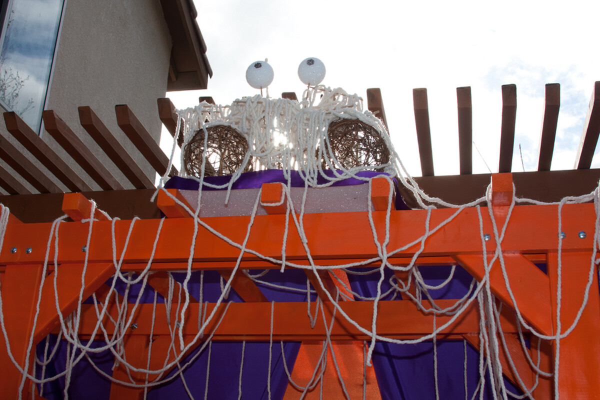 A large Flying Spaghetti Monster sculpture mounted above an orange-and-purple painted wooden arbor at a wedding reception &mdash; two wicker-ball eyes on stalks, white string noodles draped everywhere, greenery and decorations around the frame.