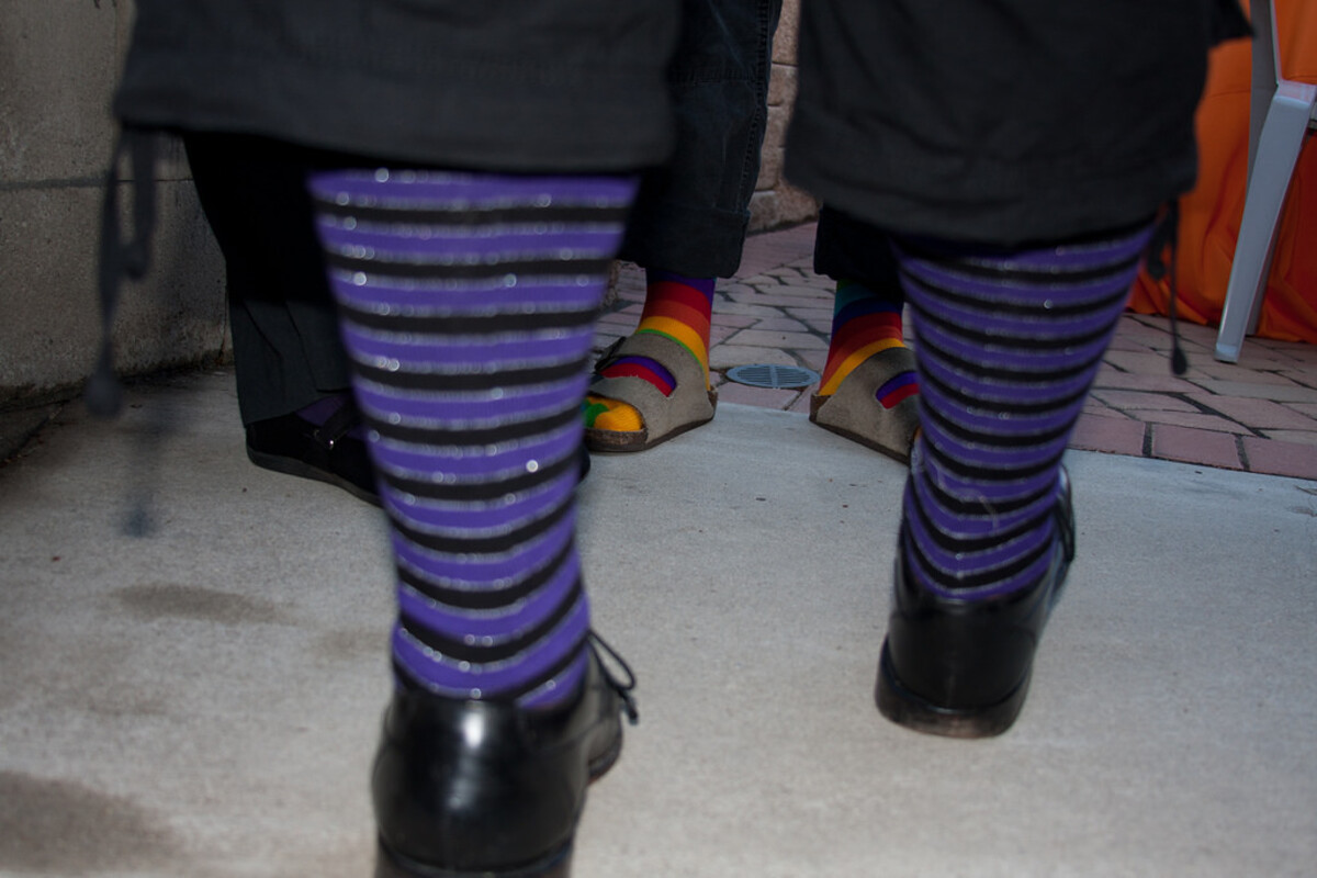 Two pairs of legs from the knees down at a wedding reception &mdash; one in purple-and-black stripy tights with black platform boots, the other in rainbow-striped socks with tan open-toed sandals, standing on a stone patio with an orange cloth-draped chair behind them.