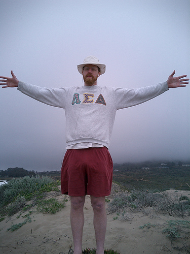 Large man with a red beard and white bucket hat, wearing a fraternity sweatshirt and red shorts, standing on sand dunes with arms spread wide against an overcast sky.