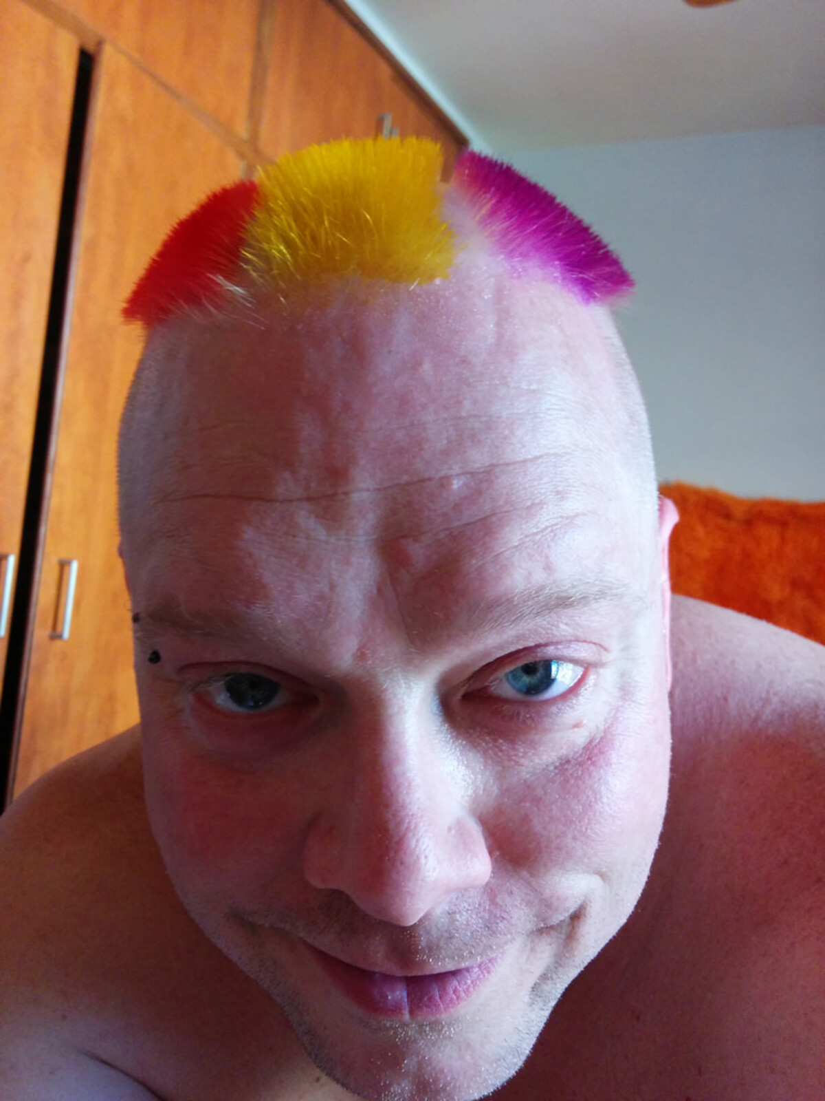Close-up selfie of Rob with a short mohawk strip in three colors: red on the left, yellow in the center, hot pink on the right. Shaved sides, bridge piercing. Taken in a bedroom.