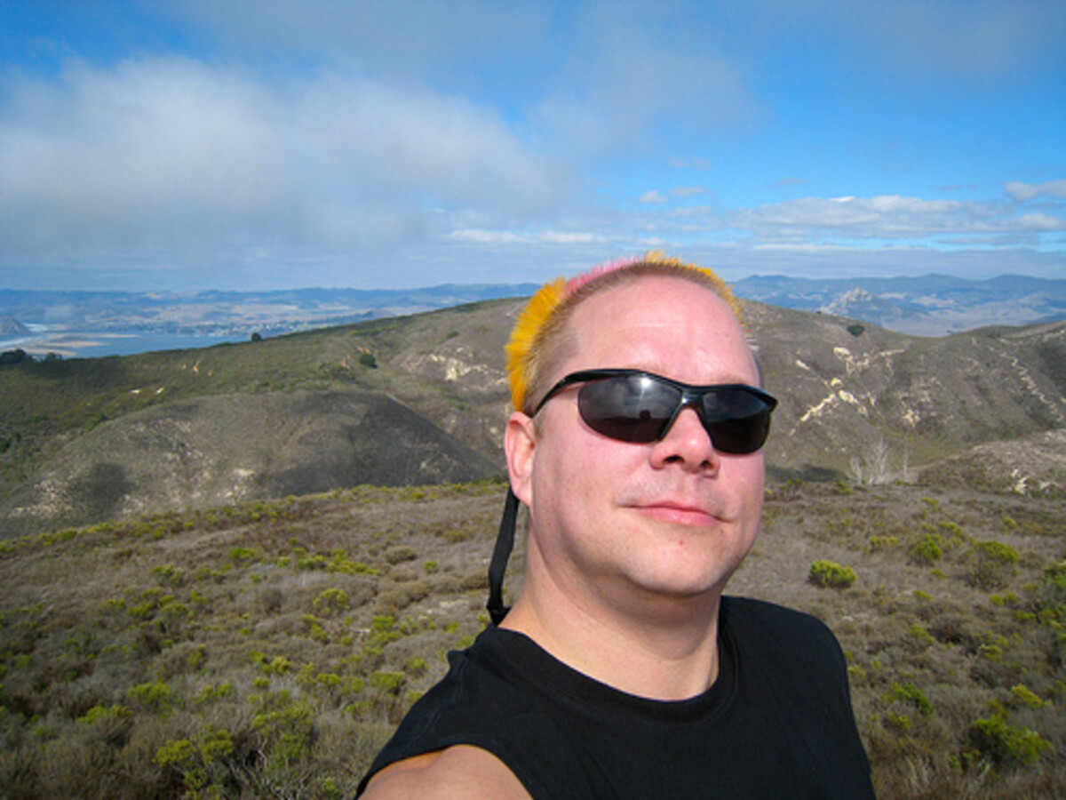 Rob taking a selfie at a hilltop at Montana de Oro, yellow-orange mohawk strip against sunlit green hills and blue sky, wearing black shirt and sunglasses.