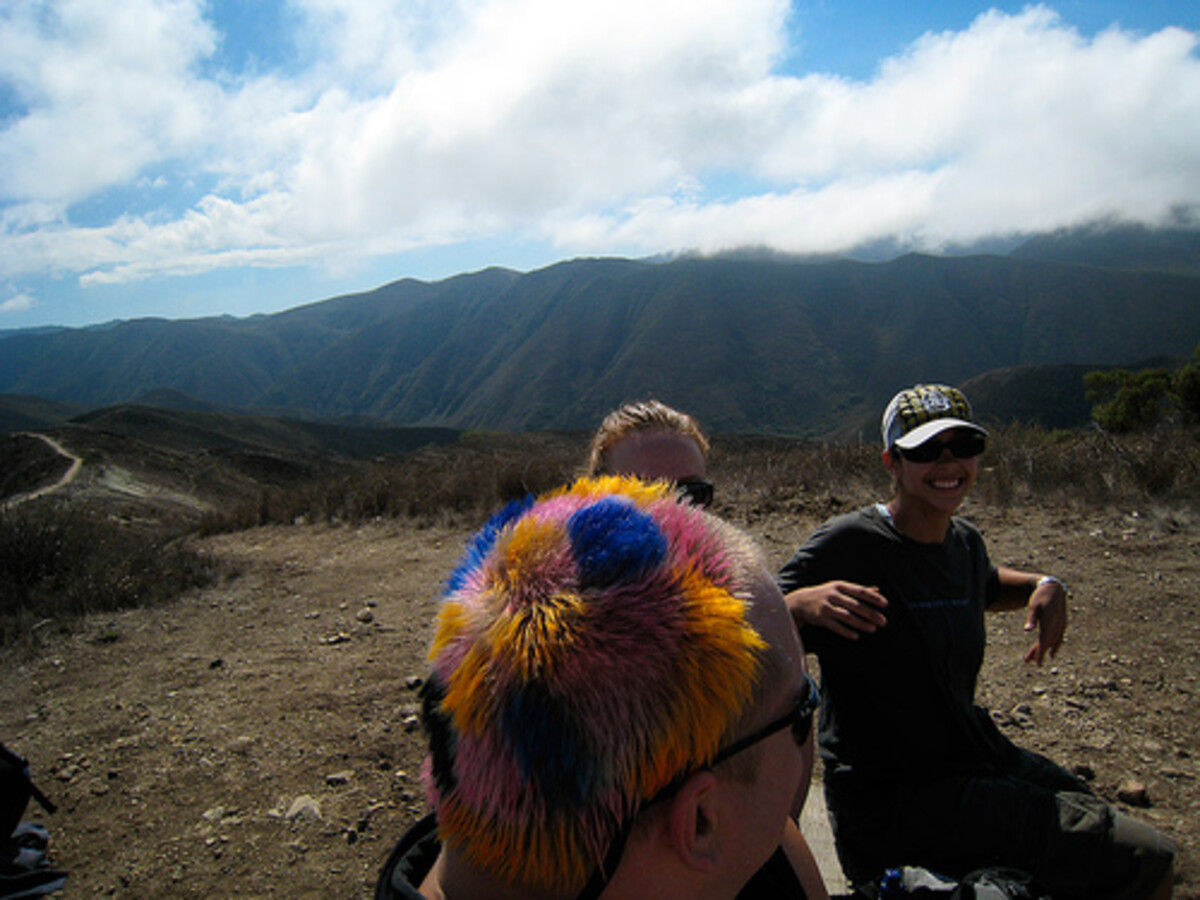 Rob on a hilltop trail wearing an enormous multicolored spiky fur hat covering his entire head, blue and orange and yellow fibers radiating outward. A smiling friend in a cap stands behind him.