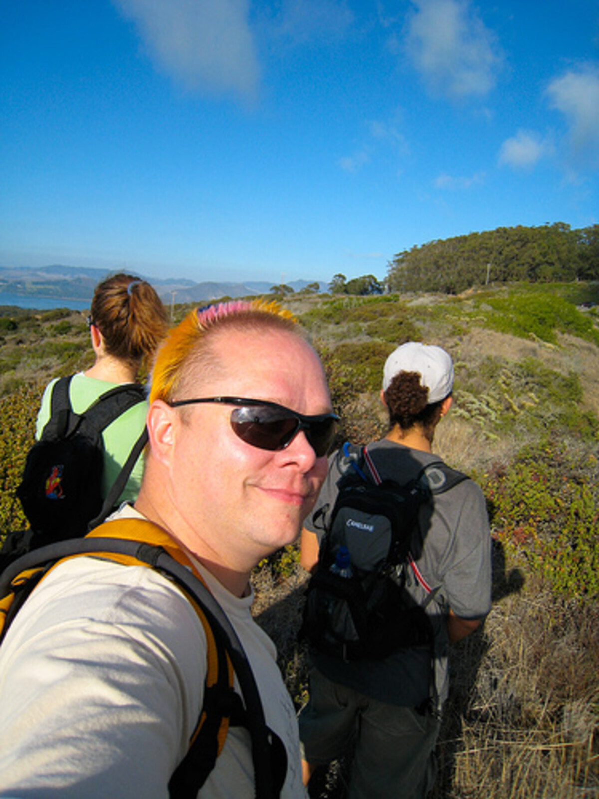 Rob hiking on a coastal trail, smiling over his shoulder at the camera, pink mohawk visible above sunglasses. Two friends with backpacks follow behind on the grassy path.