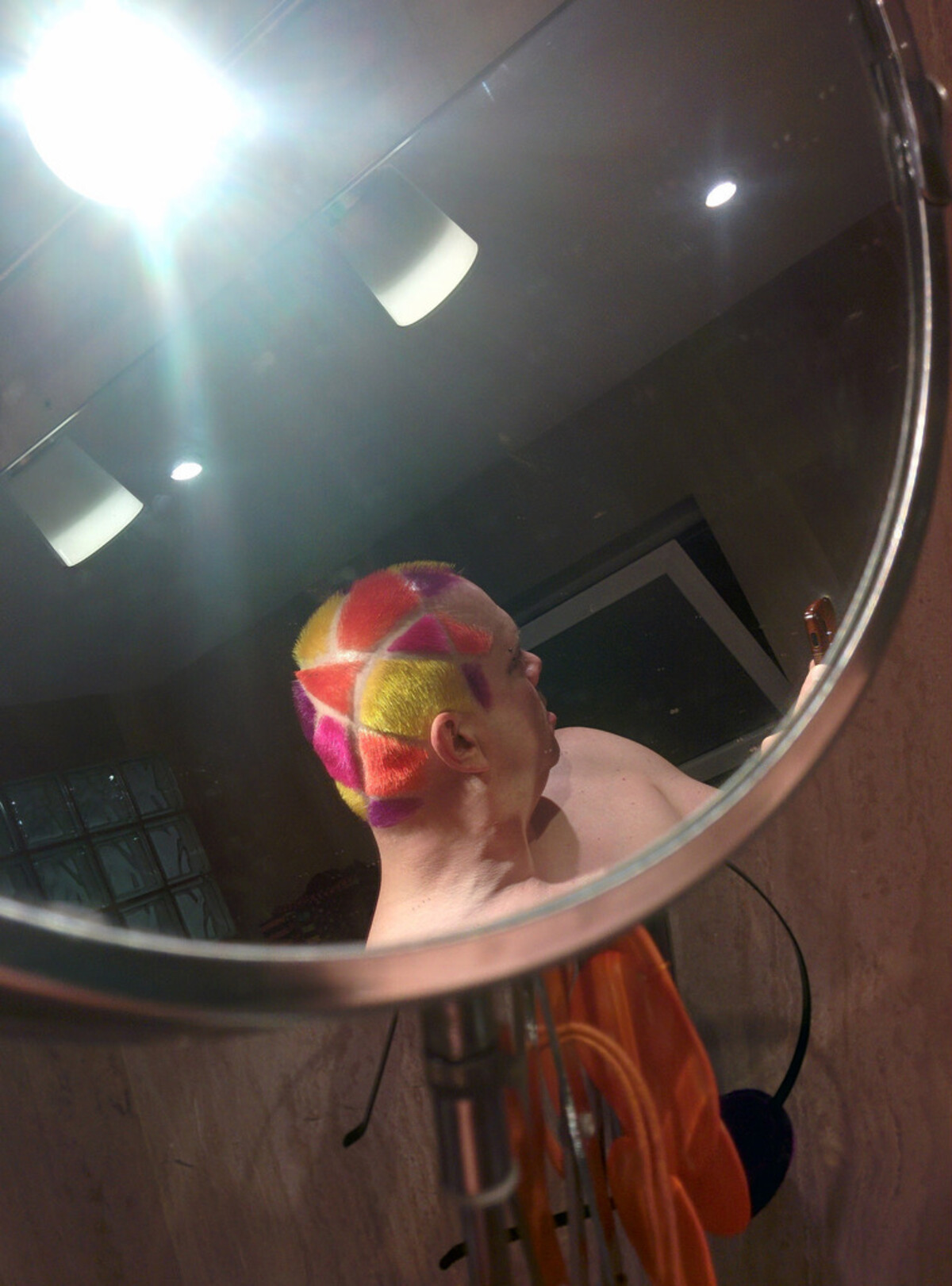 Rob reflected in a round bathroom mirror, head tilted back to show a geometric stained-glass pattern on his scalp: triangular segments in hot pink, coral, yellow, and magenta separated by shaved skin lines. Shower tiles and an orange towel visible.
