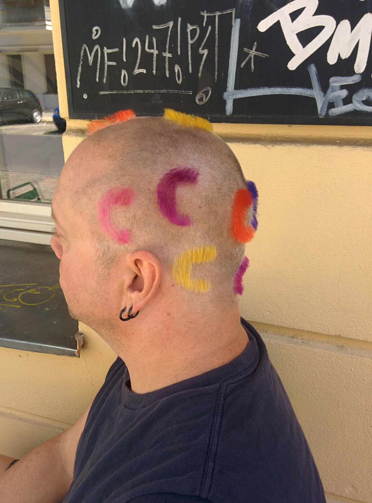 Side view of Rob's mostly shaved head with scattered crescent-shaped tufts of dyed hair in pink, magenta, purple, yellow, orange, and blue, like confetti. Black hoop earring. Chalkboard menu in background.