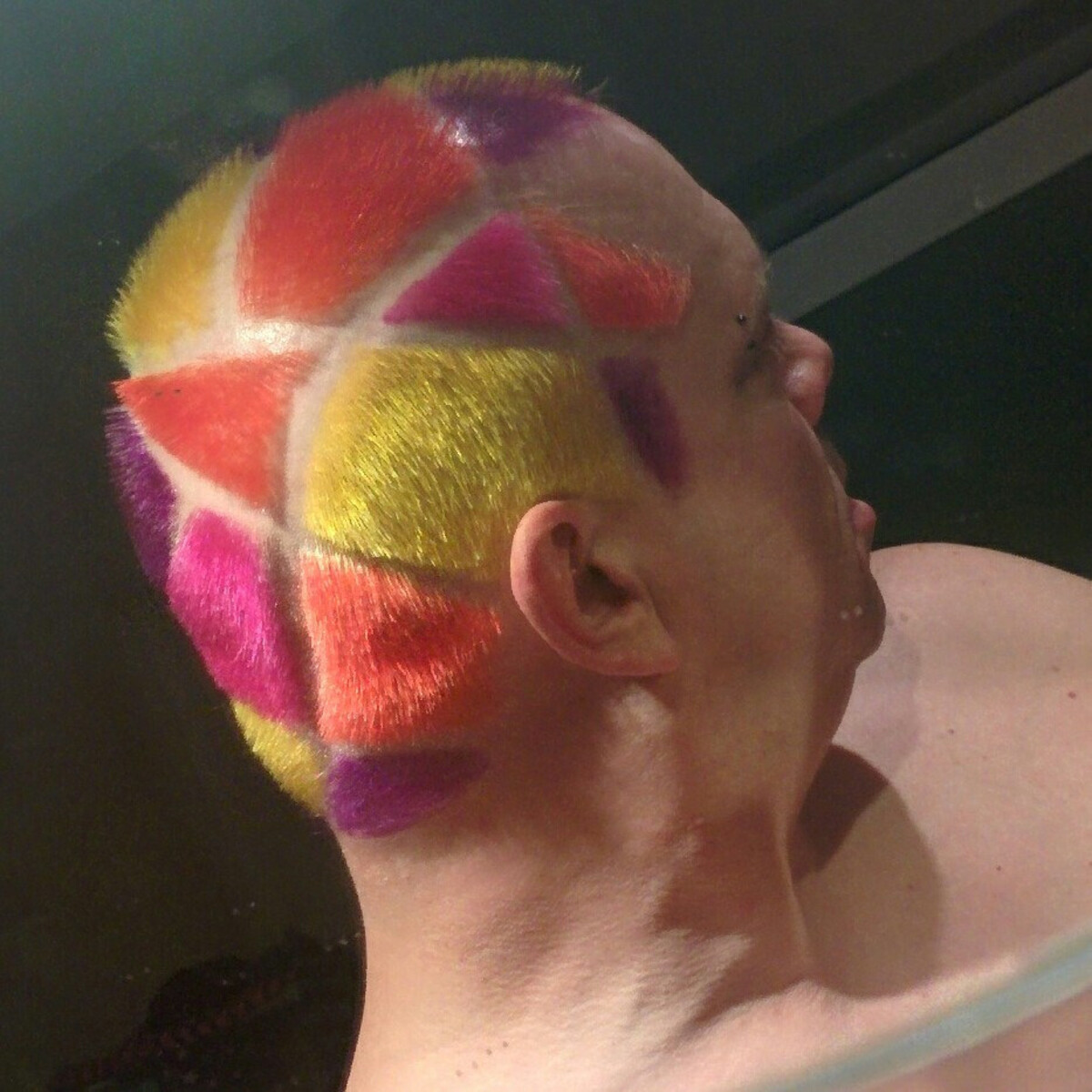 Rob shot from below showing the geometric stained-glass hair pattern in full: large triangular segments of neon yellow, coral, hot pink, and deep magenta radiating from center, with shaved lines between. Bridge piercing visible.